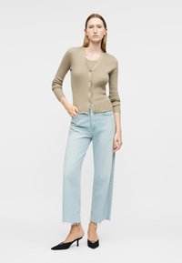 Γυναίκα με μπεζ πλεκτή ζακέτα με κουμπιά, ανοιχτό μπλε κομμένα τζιν με ξέφτια στο τελείωμα και μαύρα μυτερά παπούτσια με τακούνι kitten, στέκεται μπροστά σε λευκό φόντο.