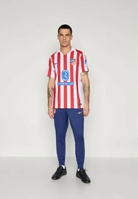 Atletico Madrid fotbaldrakt har røde og hvite vertikale striper, blå detaljer, med marine joggebukser og svarte fotballsko.