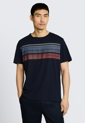 Mężczyzna noszący granatowy t-shirt z poziomymi paskami w odcieniach szarości, niebieskiego i czerwonego na wysokości klatki piersiowej, ręce w kieszeniach.