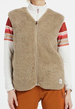 Veste beige duveteuse à fermeture éclair portée par-dessus un haut blanc à manches longues avec des rayures multicolores sur les manches et un pantalon couleur rouille.