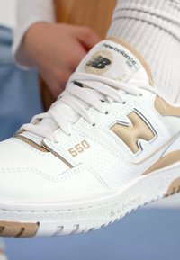 Weiße Ledersneaker mit goldenen Akzenten, ausgestattet mit einer perforierten Textur, Schnürsenkeln und der Zahl "550", die an der Seite hervorgehoben ist.