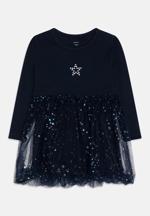 Granatowa sukienka z długim rękawem dla malucha, z kryształową gwiazdą na piersi i nakładką na spódnicę ozdobioną holograficznymi wzorami gwiazd.