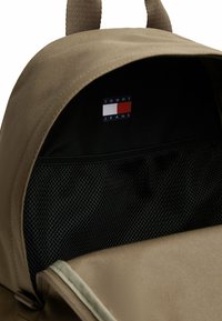 Interior de una mochila de tela marrón con un bolsillo de malla negra y una etiqueta con el logotipo "TOMMY JEANS" en colores rojo, blanco y azul.