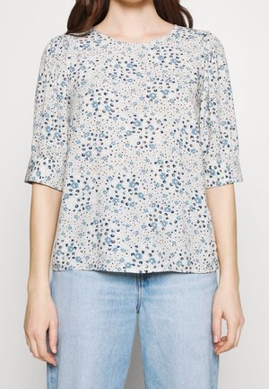 Femme portant un chemisier gris clair avec de petits motifs floraux bleus et des manches trois-quarts, associé à un jean bleu clair.
