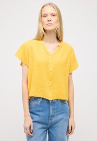Gul, tekstureret, kortærmet skjorte med knapper og V-udskæring i en afslappet pasform, parret med lys blå højtaljede jeans.