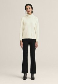 Kermainen pooloneule yhdistettynä mustiin liehuviin housuihin. Neuleessa on resorit hihansuissa ja helmassa; mallilla on jalassaan mustat nilkkurit.