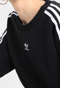 Černé tričko s krátkými rukávy, s bílými třípruhovými detaily na ramenech a malým bílým logem Adidas na hrudi.