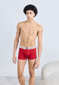 Κόκκινα βαμβακερά μποξεράκια με λευκή μέση που φέρει το λογότυπο και το έμβλημα "TOMMY HILFIGER". Το μοντέλο στέκεται σε όρθια στάση.