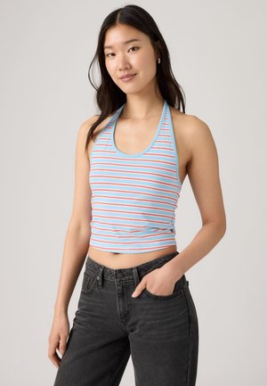 Jeune femme portant un top à bretelles rayé bleu clair et rouge et un jean noir taille haute, une main dans la poche.