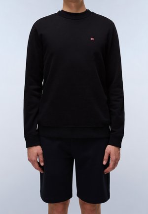 Person trägt ein schlichtes schwarzes Langarm-Sweatshirt mit einem kleinen rot-weißen Flaggenemblem auf der Brust und schwarze Shorts.