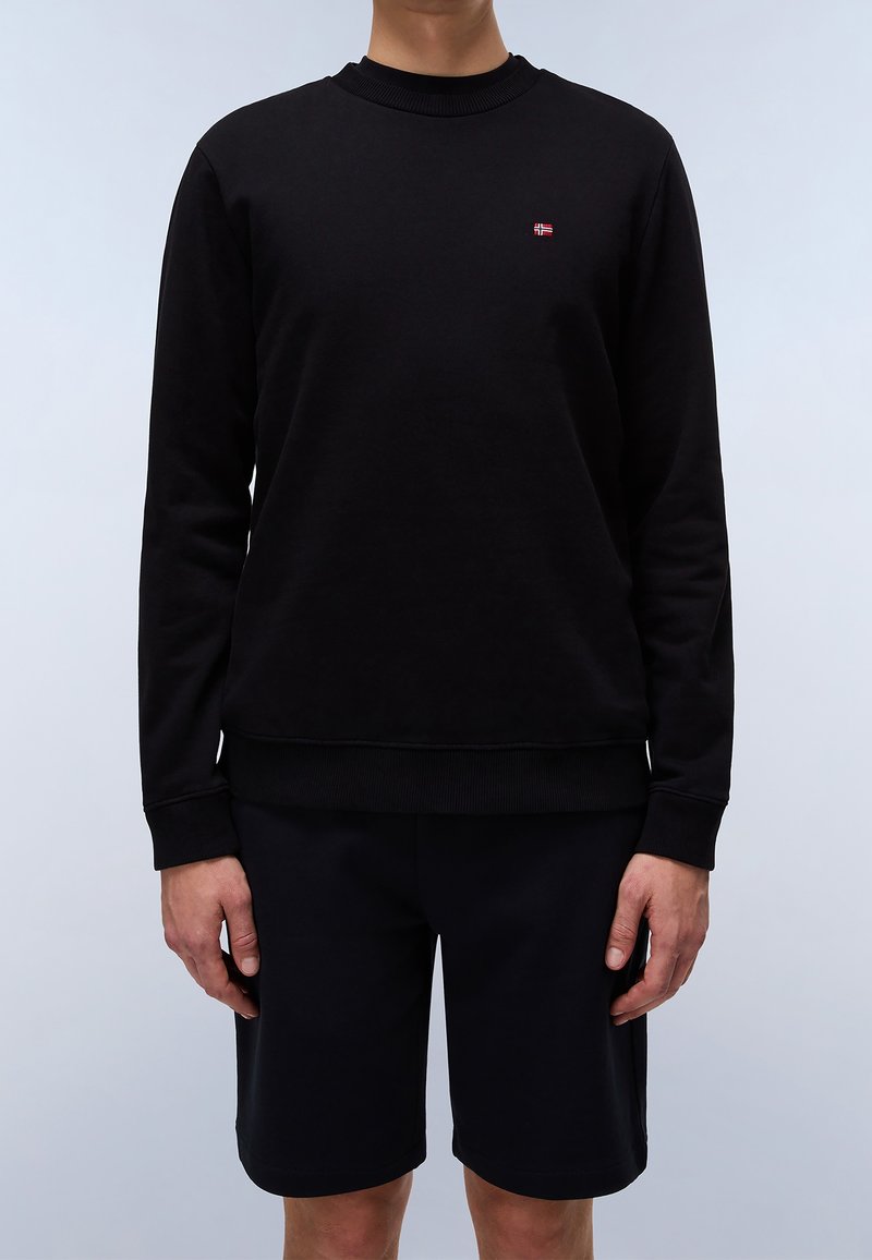 Personne portant un sweat-shirt noir uni à manches longues avec un petit emblème de drapeau rouge et blanc sur la poitrine et un short noir.