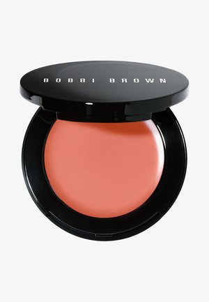 Blush crème dans un compact noir. Le produit est d'une teinte corail pêche avec une texture lisse. Le compact a une finition brillante et un logo dessus.