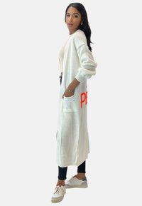Larga chaqueta blanca con textura acanalada, que cuenta con profundos bolsillos y un texto impreso en brillante color naranja. Llevada sobre una blusa ajustada y leggings negros.