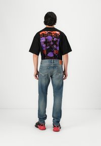 T-shirt nera oversize con stampa grafica vivace sul retro, abbinata a jeans blu sbiaditi e sneakers a fantasia rossa e nera.