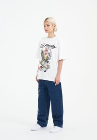 Vit oversized t-shirt med färggrafisk design, som visar ett skalle, en örn och text. Stylar med blå denimbyxor i vid modell och vita sneakers.