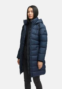 Mujer vestida con un abrigo acolchado largo y azul marino con capucha, una blusa negra y jeans oscuros, de pie contra un fondo claro y liso.
