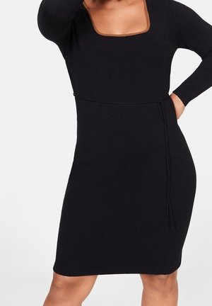 Femme portant une robe noire ajustée à manches longues, avec un col carré et une fine ceinture nouée à la taille.