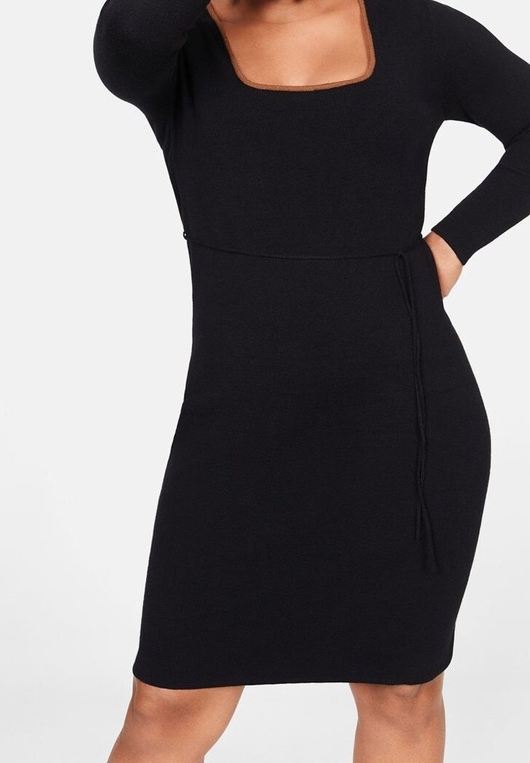 Femme portant une robe noire ajustée à manches longues, avec un col carré et une fine ceinture nouée à la taille.