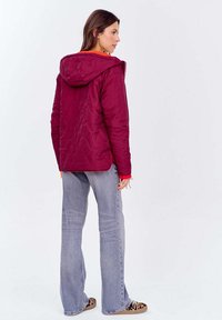 Kobieta w bordo kapturze, w pikowanej kurtce, jasnoniebieskich dżinsach i trampkach w panterkę stoi zwrócona na prawo na białym tle.