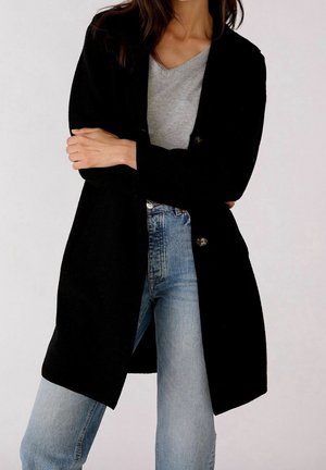 Femme portant un cardigan noir boutonné, un t-shirt gris à col en V et un jean bleu clair, debout les bras croisés devant un fond uni.