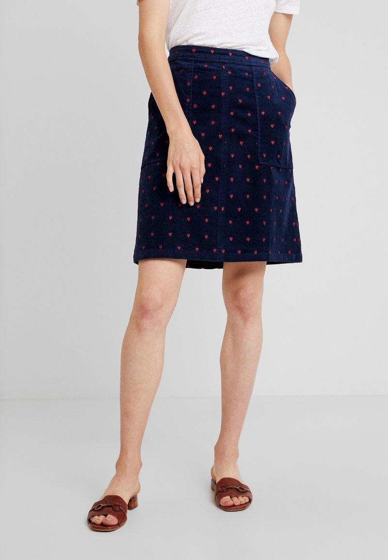 Femme portant une jupe bleu marine avec de petits cœurs rouges, un haut blanc et des sandales ouvertes marron, debout avec une main dans la poche.