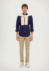 Marineblå skjorte med lange ermer, kremfarget strikkegenser knyttet rundt nakken, khakibukser og hvite sneakers med svarte detaljer.