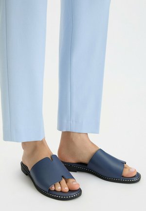Füße tragen marineblaue offene Slipper-Sandalen mit Einkerbung, kombiniert mit hellblauen gekürzten Hosen auf weißem Hintergrund.