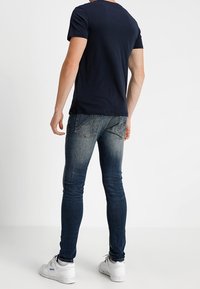 Man som bär en mörkblå t-shirt, bleka skinny jeans och vita sneakers, som står vänd bort mot en vit enkel bakgrund.