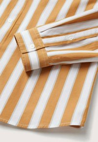 Camicia a righe giallo senape e bianca. Tessuto in cotone con polsini abbottonati e righe verticali distintive di larghezze variabili.