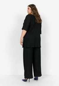 Tunique noire texturée à manches courtes et fentes latérales, associée à un pantalon noir ample. Le modèle porte des chaussures à talons hauts bleues.