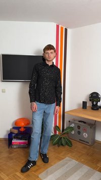 Camicia nera con bottoni e macchie bianche, jeans azzurri chiari e sneakers nere. Ambientazione interna con strisce colorate sulle pareti e mobili.
