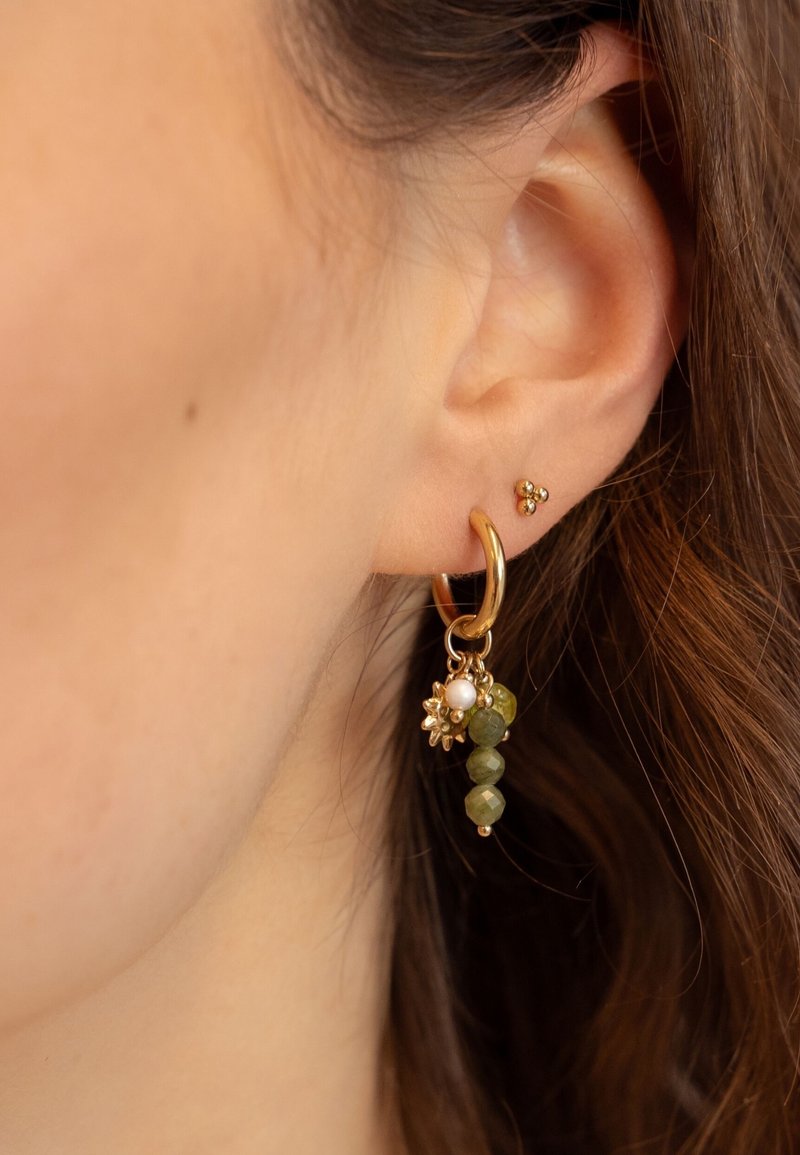 Pendiente de aro dorado con piedras preciosas verdes, acentos florales y una perla; se lleva junto a un pequeño pendiente de botón en la oreja.