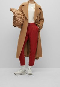 Abrigo largo color camel sobre una camisa crema con "BOSS" en dorado, combinado con pantalones rojos y zapatillas ligeras de color gris. Se incluye un bolso acolchado marrón.