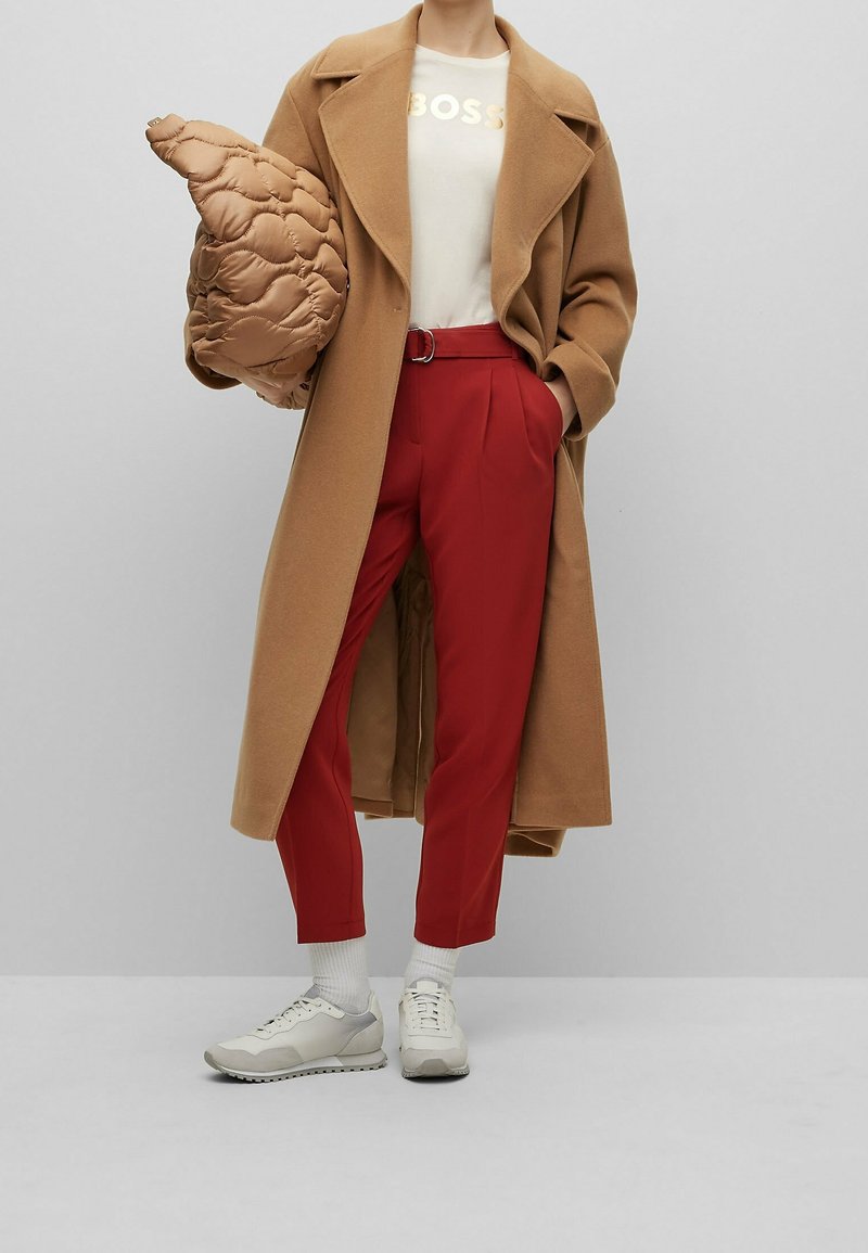 Abrigo largo color camel sobre una camisa crema con "BOSS" en dorado, combinado con pantalones rojos y zapatillas ligeras de color gris. Se incluye un bolso acolchado marrón.