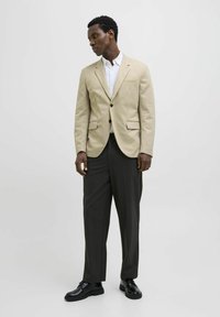 Beige blazer with a single pocket, paired with a white shirt and dark trousers. Black shoes complete the outfit. Simple, tailored design.