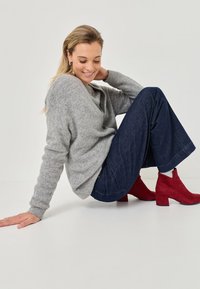 Pulover gri, confortabil, blugi larghi negri, și cizme scurte roșii din suede cu toc bloc, așezată cu o postură relaxată pe un fundal deschis.