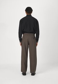 Zwarte overhemddas met lange mouwen en een losse pasvorm, gecombineerd met bruine wijde broek en zwarte schoenen, bekeken van achteren.