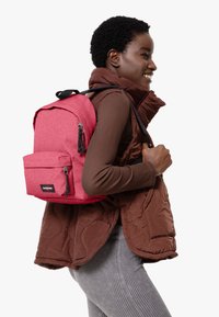 Mochila de tela rosa con un acabado texturizado, que cuenta con un bolsillo frontal y herrajes negros, llevada por una persona con un chaleco acolchado marrón.