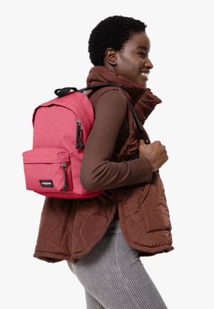 Mochila de tela rosa con un acabado texturizado, que cuenta con un bolsillo frontal y herrajes negros, llevada por una persona con un chaleco acolchado marrón.