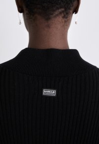 Suéter preto canelado com gola alta. Apresenta um rótulo "Barbour International" na parte de trás. Textura tricotada, design minimalista.