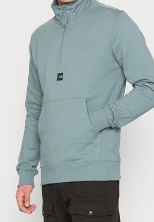 Mand iført lyseblå sweatshirt med kvart-lynlås og forlommer samt sorte cargobukser, hånd i lommen.