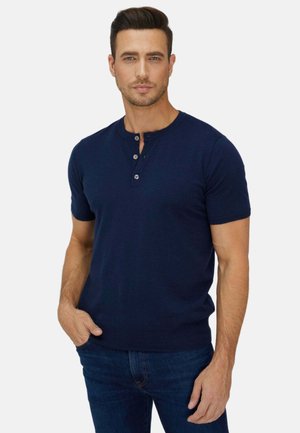 Mann mit kurzen dunklen Haaren, der ein marineblaues Henley-Shirt mit kurzen Ärmeln und blaue Jeans trägt, steht mit einer Hand in der Tasche.
