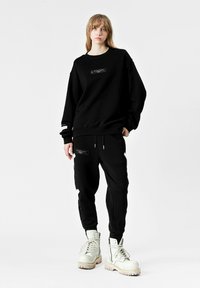 Sudadera negra de gran tamaño con un bolsillo frontal y dos rayas blancas en las mangas. Combinada con pantalones joggers negros que cuentan con bolsillos con cremallera y un diseño desgastado.