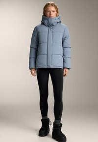 Světle modrá péřová bunda s vysokým límcem, horizontálním prošíváním, v kombinaci s černými legínami a černými šněrovacími botami. Hladká textura.