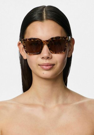 Donna con capelli lisci e scuri che indossa grandi occhiali da sole quadrati con montatura tartarugata, rivolta in avanti con un'espressione neutra su uno sfondo bianco.