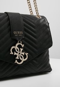 Sac à main en cuir matelassé noir Guess avec sangle chaîne dorée et pendentif logo Guess doré entrelacé sur le rabat avant.