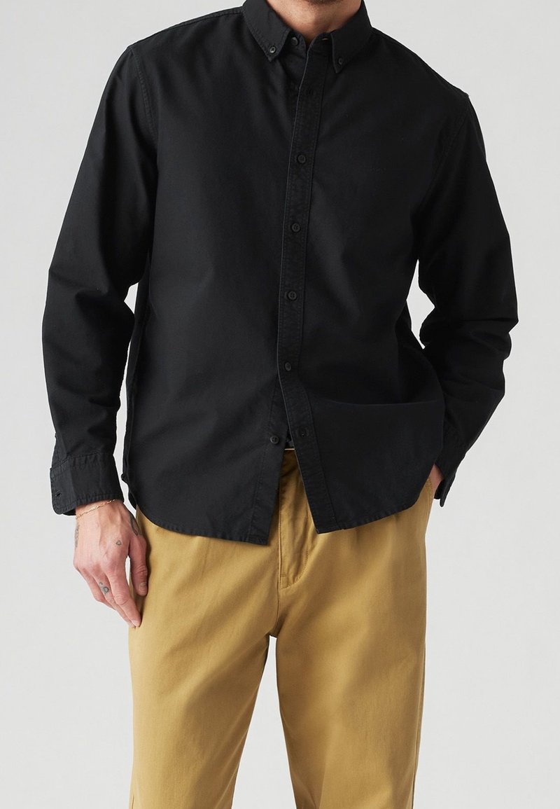 Homme portant une chemise noire à manches longues boutonnée et un pantalon jaune moutarde, debout devant un fond clair uni.