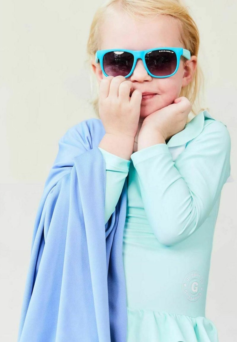 Aquamarine long-sleeve swimsuit with a ruffled hem, paired with a light blue cover-up. Child wears turquoise sunglasses, smiling with hands to face.