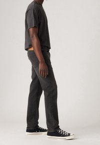 Mörkgrå jeans med raka ben, kombinerade med en mörk t-shirt. Skorna är svarta tygskor med vita sulor och snören.