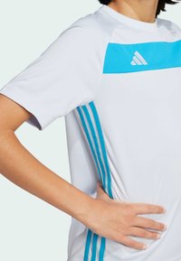 Camiseta deportiva blanca con una franja horizontal turquesa y tres franjas verticales turquesas a cada lado. Cuenta con cuello redondo y corte estándar.